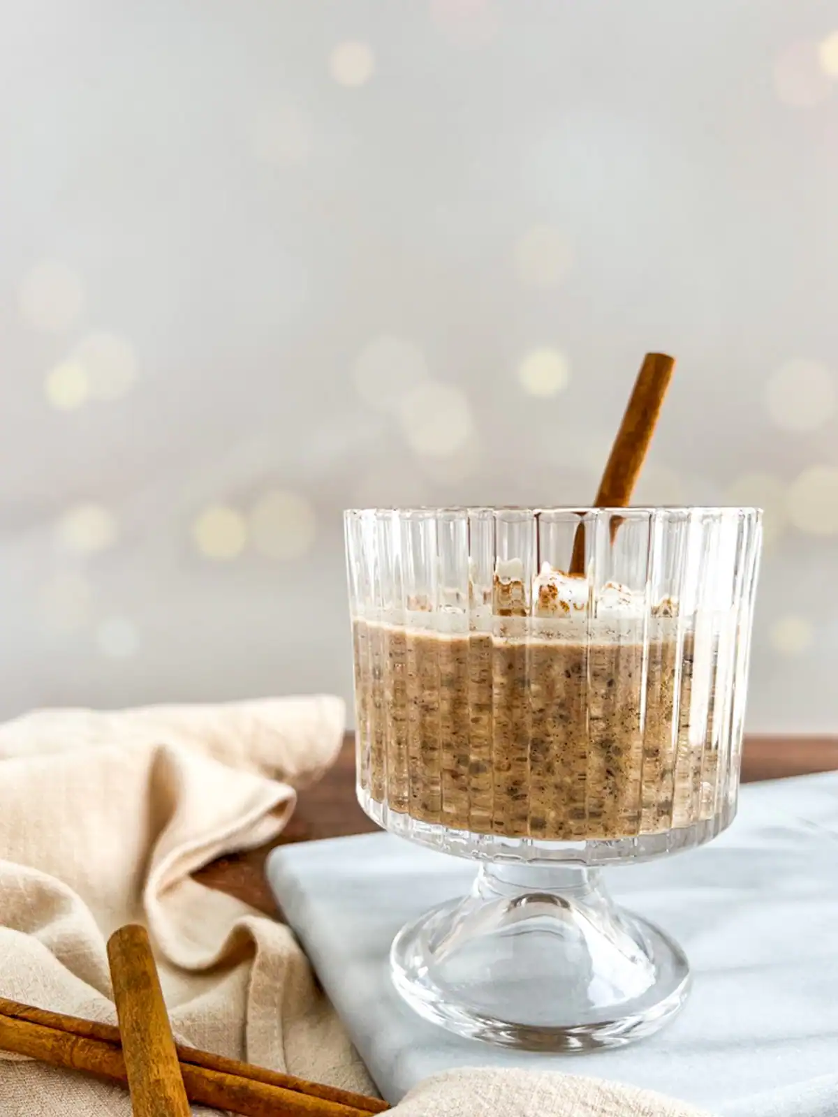 Chai spice overnight oats in a textured glass with cinnamon sticks, served on a light cloth and marble surface.