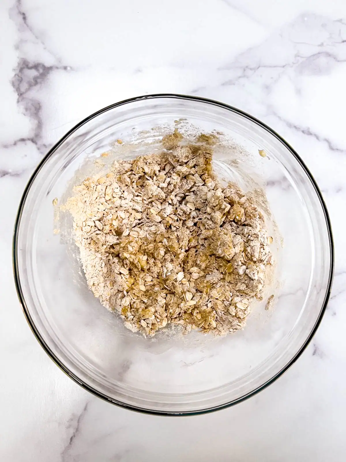 Mashed banana mixed with oats in a glass mixing bowl on marble surface.