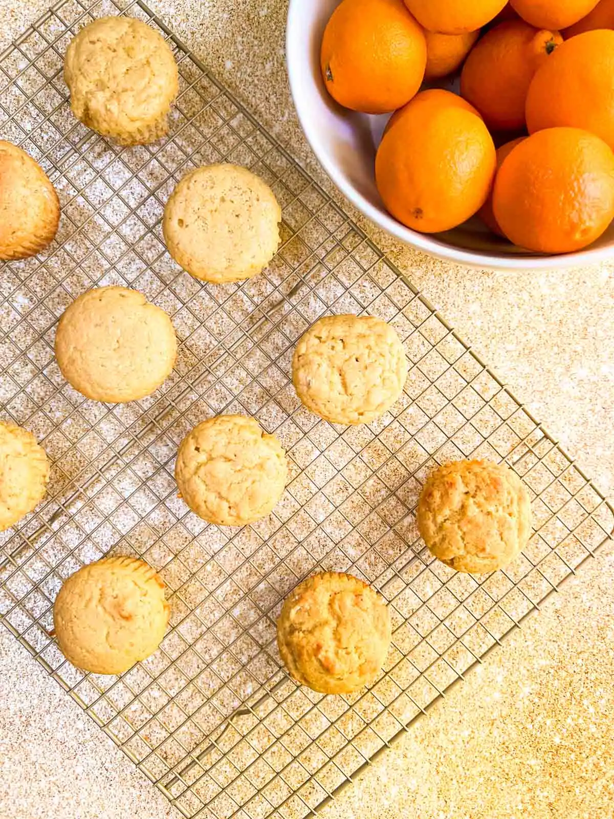 Vegan orange vanilla muffins cooling on a wire rack with a bowl of fresh oranges nearby.