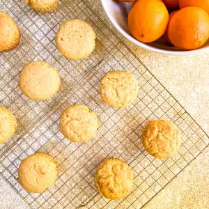 Freshly baked vegan orange muffins on a cooling rack with a bowl of oranges in the background.