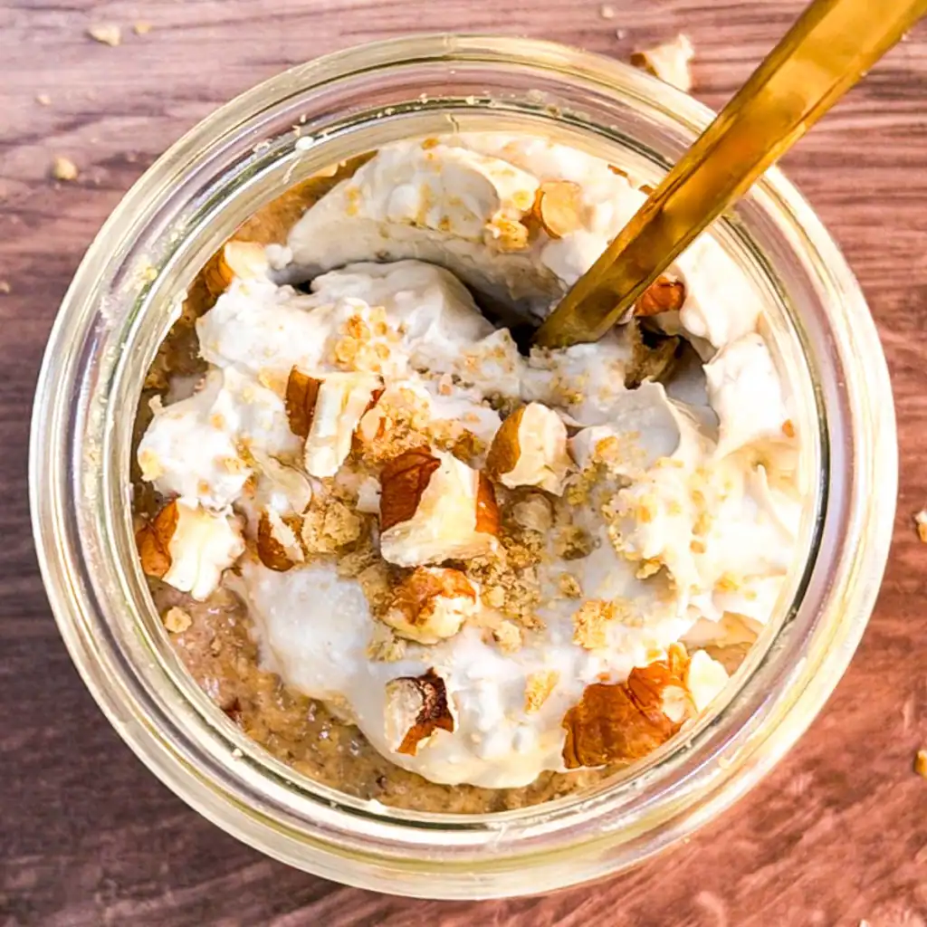 Blended vegan pumpkin spice overnight oats in a glass jar topped with crushed nuts and coconut whipped cream.