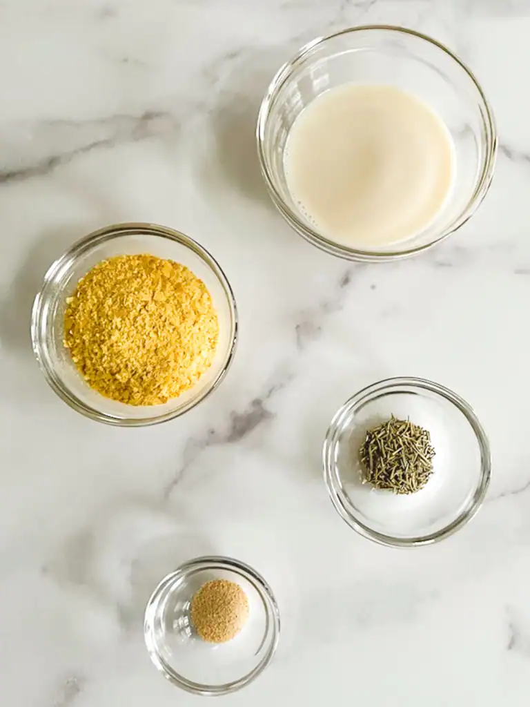 Creamy plant-based milk, nutritional yeast, herbs, and seasonings in glass bowls on white marble surface, for vegan mashed potatoes.