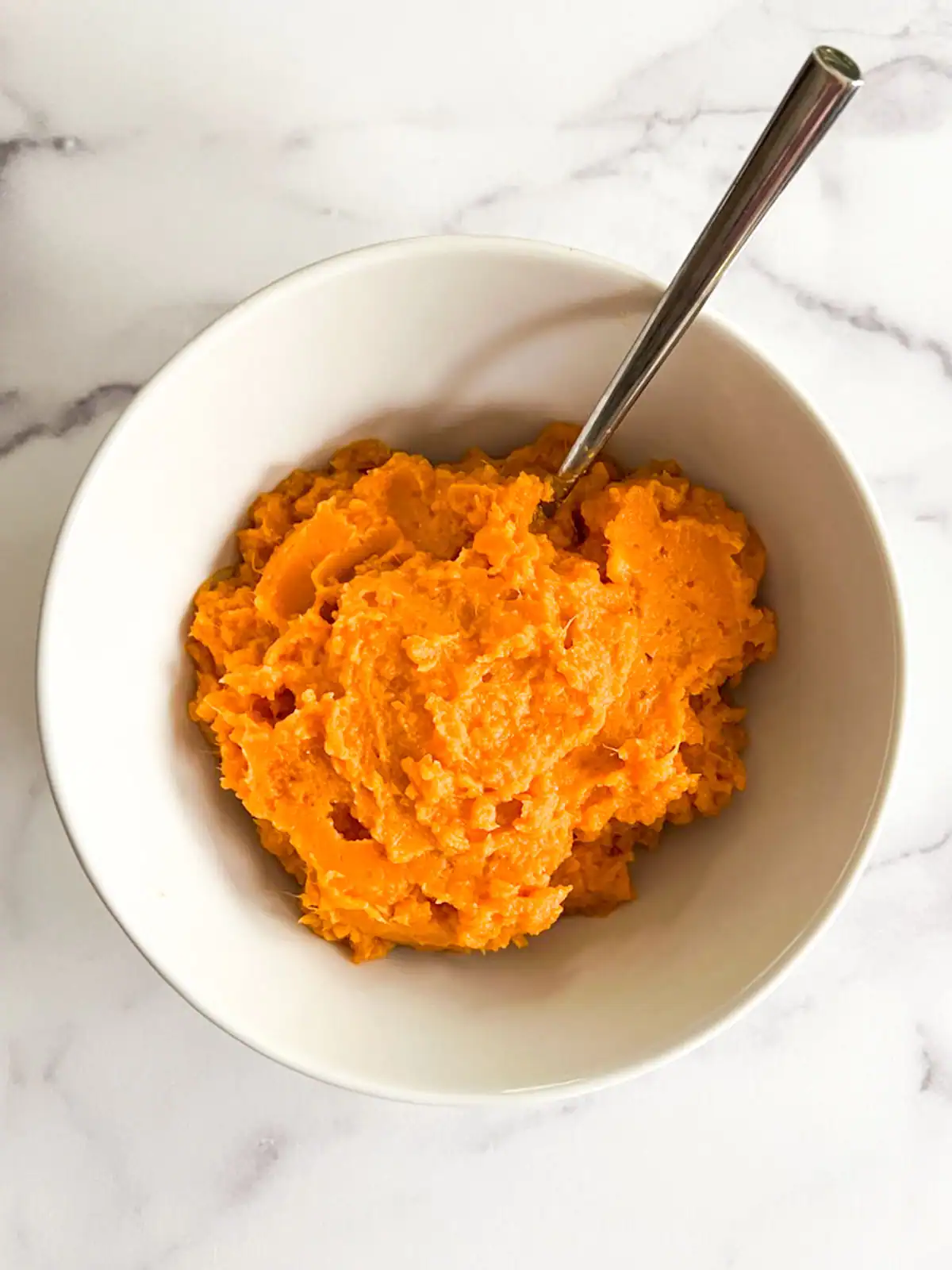 Creamy mashed sweet potatoes in a white bowl with a metal spoon.