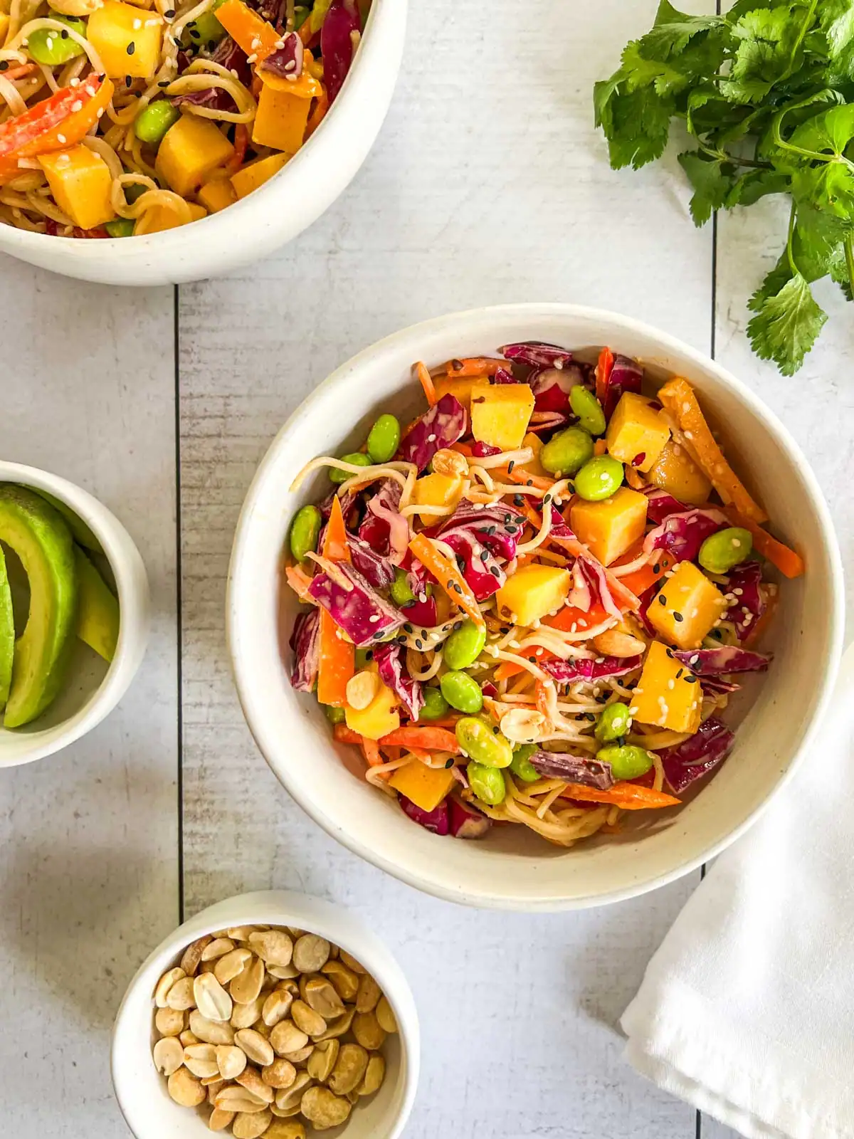 Mango peanut noodle salad in a white bowl on a table surrounded by garnishes.