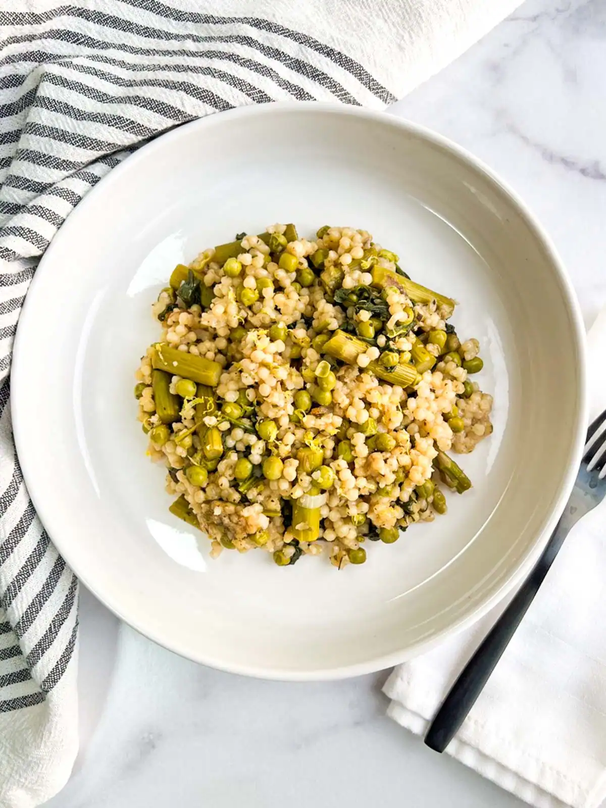 Baked pearl couscous with asparagus and peas on a white plate next to a striped cloth.