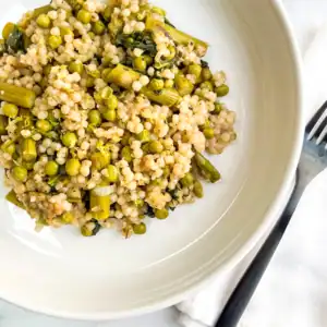 Baked pearl couscous with asparagus. and peas on a white plate.
