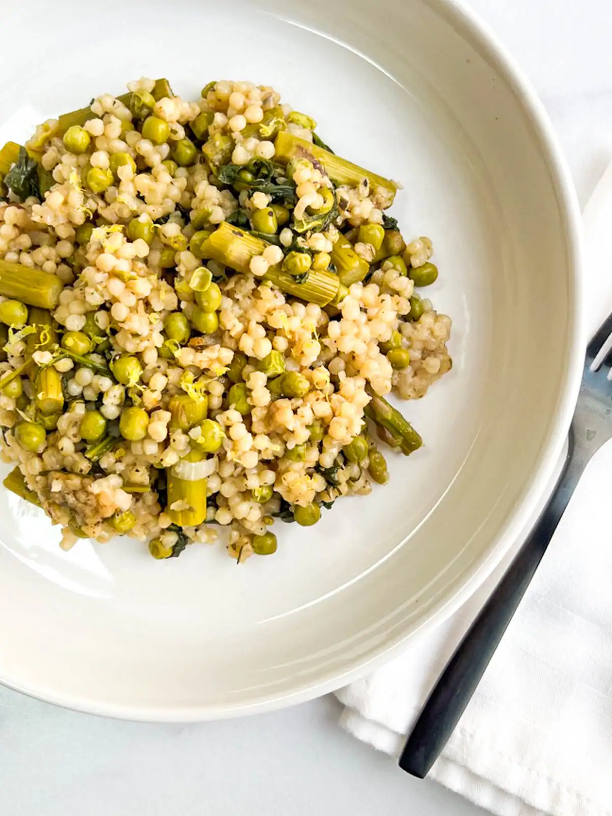 Baked pearl couscous with asparagus. and peas on a white plate.