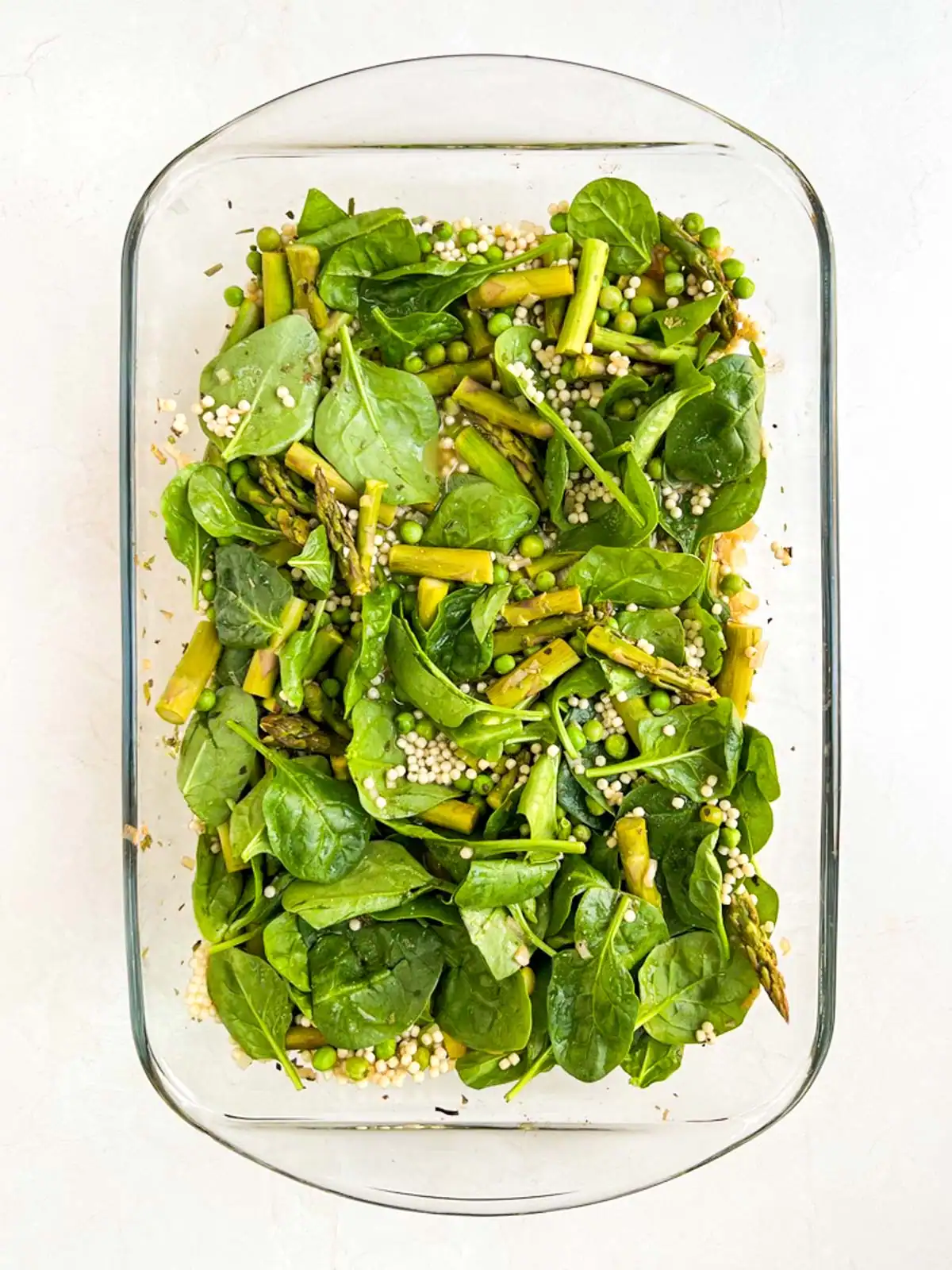 Spinach and pearl couscous added to ingredients in glass casserole dish.