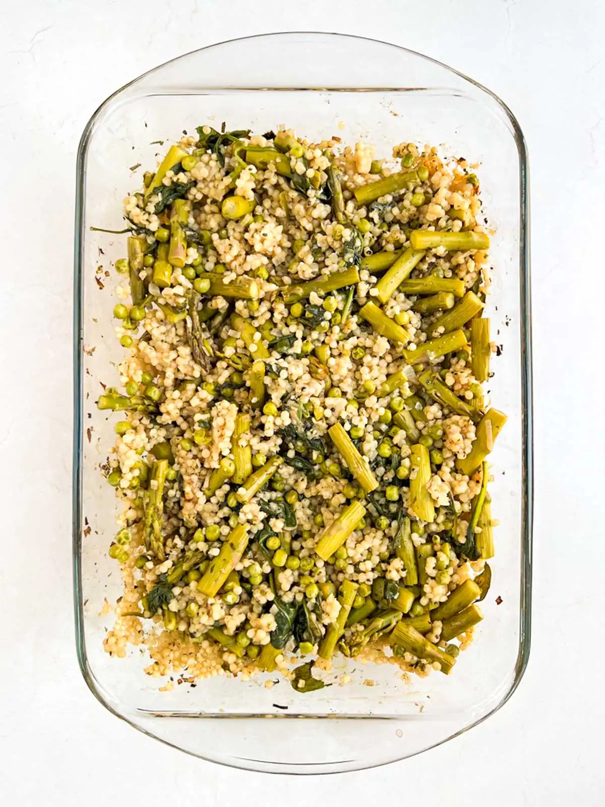 Baked pearl couscous with asparagus and peas in a glass casserole dish.